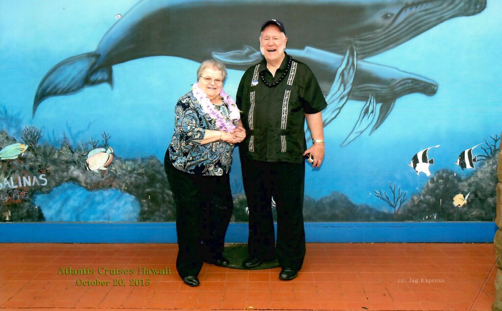 Dr. Jerry and Elaine Ramsey, wearing leis, pose on a Hawaiian cruise in front of a mural painted with whales.